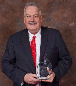 Steve Fronek, wearing a black suit, white dress shirt and red tie, holds his Architectural Distinguished Service Award and smiles for the camera.