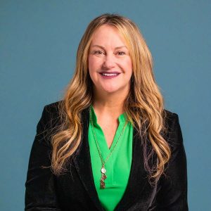 Jaclyn Harrison, wearing a green button up shit, black jacket, and longer gold necklace, stands in front of a blue screen and smiles for the camera.